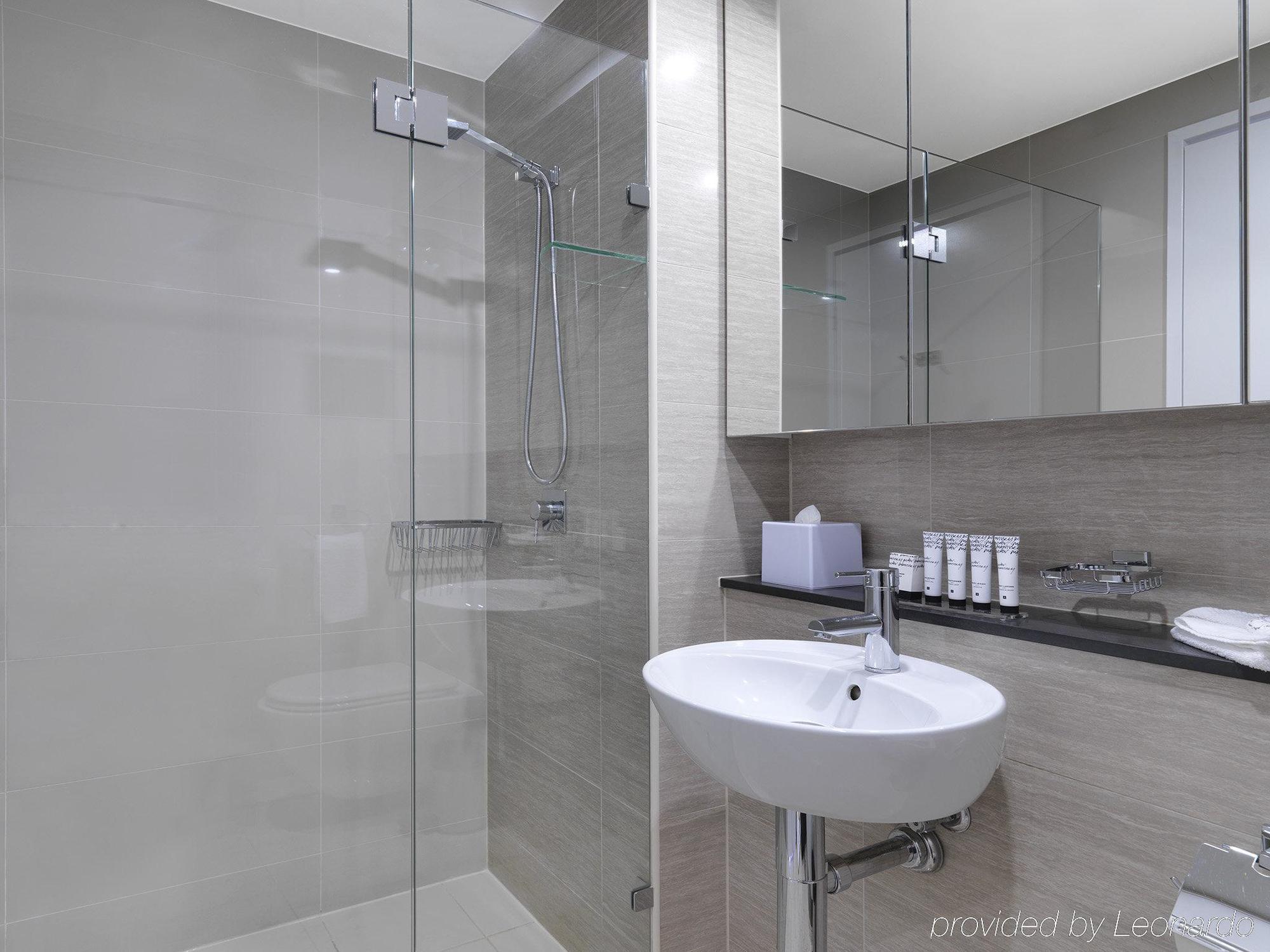 Meriton Suites Campbell Street, Sydney Exterior photo The photo shows a modern bathroom featuring a glass shower enclosure with a rainfall showerhead. There is a sleek white basin with a contemporary faucet mounted on a dark countertop. The walls are finished in light gray tiles, and there are mirrors t